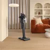 A cordless vacuum cleaner is standing on a wooden floor in a living room, with a sofa and shelves in the background. Anteprima dell’immagine prodotto