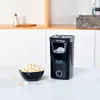 A black popcorn maker sits on a white countertop next to a bowl of popcorn. Produktbild-Vorschau