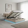A bedroom with a beige upholstered bed frame that has its storage compartment open, revealing storage boxes and pillows. The bed has LED lighting around its base. Produktbild-Vorschau