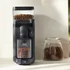 A black coffee grinder with coffee beans in the hopper and ground coffee in the collection bin, placed on a countertop next to a glass jar filled with coffee beans. Produktbild-Vorschau