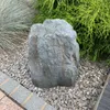 A decorative stone cover in a garden setting, surrounded by gravel and plants. Produktbild-Vorschau
