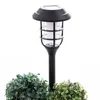 A black solar-powered garden light stands between two artificial green bushes against a white background. Produktbild-Vorschau