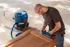 A man is using a Bosch cordless planer on a wooden door, with a Bosch vacuum cleaner connected to it. Podgląd zdjęcia produktu