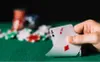 A hand holding two aces of spades and diamonds over a poker table with chips in the background. Produktbild-Vorschau