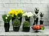 A collection of decorative items including vases with flowers, potted plants, and a fruit bowl, arranged on a surface against a brick wall. Produktbild-Vorschau