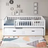 A white wooden daybed with a pull-out trundle and two drawers, set against a light gray wall with decorative letters and framed pictures. Produktbild-Vorschau