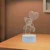 A 3D wireframe teddy bear lamp with a heart-shaped balloon sits on a wooden surface against a brown background. Produktbild-Vorschau