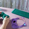 A person's hands are holding a teal game controller on a pink surface, next to a teal keyboard and purple headphones with cat ears. Náhľad obrázku produktu