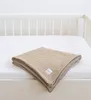 A beige muslin blanket folded on a white surface, with the edge of a white crib in the background. Produktbild-Vorschau