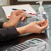 A person is applying a label that says 'Please sanitize your hands' to a bottle of hand sanitizer. Produktbild-Vorschau