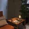 A vintage-style desk lamp sits on a wooden side table next to a chair and a large potted plant. Produktbild-Vorschau