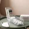 A clear organizer box filled with cables and a remote control, placed on a green table next to a white speaker and a smartphone. Produktbild-Vorschau