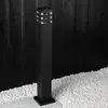 A black garden bollard light stands on a gravel and concrete surface in front of a black wooden fence. Náhľad obrázku produktu