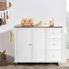 A white kitchen island with a stainless steel countertop is shown against a white wall. The island has two cabinet doors and three drawers on the right side. Various kitchen items are placed on top of the island, including a toaster, a donut on a tray, and Náhľad obrázku produktu