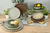 A collection of stoneware plates and bowls in various earthy tones, arranged on a wooden surface with decorative greenery and stone elements. Produktbild-Vorschau