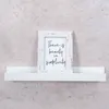 A white shelf with a framed picture on it, against a textured gray wall. Produktbild-Vorschau