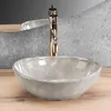 A close-up shot of a marble-patterned vessel sink with a gold faucet, set on a wooden surface with a textured gray background. Podgląd zdjęcia produktu