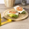 Two small open-faced sandwiches with lettuce, cheese, and tomato slices are arranged on a round wooden cutting board. A basket and a spice grinder are visible in the background, and a bottle of oil is in the foreground. Produktbild-Vorschau