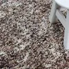 A close-up view of a shaggy rug with a speckled pattern of beige, brown, and white fibers. A portion of a white stool or table leg is visible on the right side of the image. Podgląd zdjęcia produktu