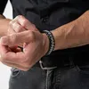 A man is adjusting his bracelet while wearing a black shirt and jeans. Anteprima dell’immagine prodotto