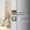 A close-up of a bronze-colored door handle set on a white door, with a blurred background of a room interior. Produktbild-Vorschau