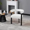 A white armchair with black metal legs is placed on a gray rug in front of a white fireplace. A coffee table with bread and a vase is to the left of the armchair. Produktbild-Vorschau