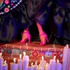 A close-up view of a doll's legs and feet wearing pink high-heeled shoes, standing on a decorated brick surface with candles in the foreground. Náhled obrázku produktu
