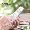 A person's hands are holding a white cordless phone, with a blurred outdoor background of greenery and a wooden table with glasses and a tablet. Anteprima dell’immagine prodotto