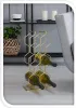 A modern living room setting with a gold wine rack holding several bottles of wine, placed on a light gray floor next to a gray armchair. In the background, a black shelf unit holds a woven basket with a plant, a white cup, and two white candles. Produktbild-Vorschau