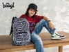 A young boy wearing a baseball cap and casual clothing is sitting on a wooden table next to a backpack. Podgląd zdjęcia produktu