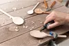 A person's hand is using a Dremel rotary tool to sand a wooden spoon on a wooden workbench. Several other wooden spoons and carving tools are visible. Anteprima dell’immagine prodotto