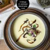 A bowl of creamy soup with garnishes, presented on a rustic plate, with ingredients like nuts and seeds in the background. Produktbild-Vorschau