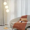 A modern floor lamp with three white glass globes stands next to a sofa with a throw blanket and pillow. Podgląd zdjęcia produktu
