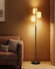 A modern floor lamp with three fabric shades stands next to a sofa in a living room. Produktbild-Vorschau
