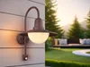 An outdoor wall light illuminates a patio area with a sofa and trees in the background. Produktbild-Vorschau