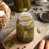 A jar of pickles with a fermentation weight on top, surrounded by ingredients like onions, carrots, bay leaves, and peppercorns, on a wooden surface. Produktbild-Vorschau