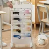 A person's hand is shown adjusting a mobile organizer cart filled with magazines and other items, placed next to a desk and chair. Produktbild-Vorschau
