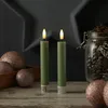 Two green LED candles with flickering flames are placed on a wooden surface, surrounded by decorative Christmas ornaments and a jar of pinecones. Produktbild-Vorschau