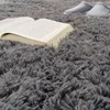 A close-up of a fluffy gray rug with an open book and a pair of slippers in the background. Voir les images du produit