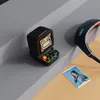 A small retro-style speaker with a pixelated screen and colorful keyboard keys sits on a gray surface next to a pair of headphones and a small photo of a person. Náhled obrázku produktu