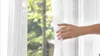 A hand is pulling back a white sheer curtain to reveal a blurred outdoor scene with trees. Produktbild-Vorschau