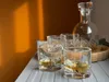 Whiskey glasses and a decanter are arranged on a shelf, with warm sunlight casting shadows on an orange wall. Produktbild-Vorschau
