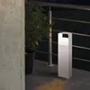 A white bollard light is shown on a patio at night, next to a potted plant and a concrete wall. Produktbild-Vorschau