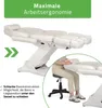 A white electric cosmetic bed with a person sitting on a stool next to it, demonstrating its use. Produktbild-Vorschau