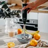 A hand holding a black immersion blender over a bowl of bananas, with oranges, a glass of juice, and a vase of greenery on a kitchen counter. Produktbild-Vorschau