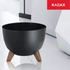 A black planter with wooden legs sits on a white marble floor, with a white geometric structure in the background. Náhled obrázku produktu
