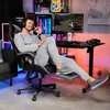 A man is sitting in a gaming chair with his feet up, in front of a computer setup. The room has ambient lighting. Produktbild-Vorschau
