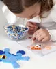 A young girl is focused on creating a perler bead design, with a bowl of beads and a partially completed blue character in the foreground. Produktbild-Vorschau
