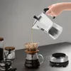 A hand pouring coffee from a moka pot into a coffee dripper over a carafe, with a coffee grinder and jar in the background and a mini hot plate in the foreground. Produktbild-Vorschau