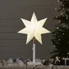 A lit paper star decoration on a stand, placed on a table with Christmas gifts and a decorated Christmas tree in the background. Podgląd zdjęcia produktu